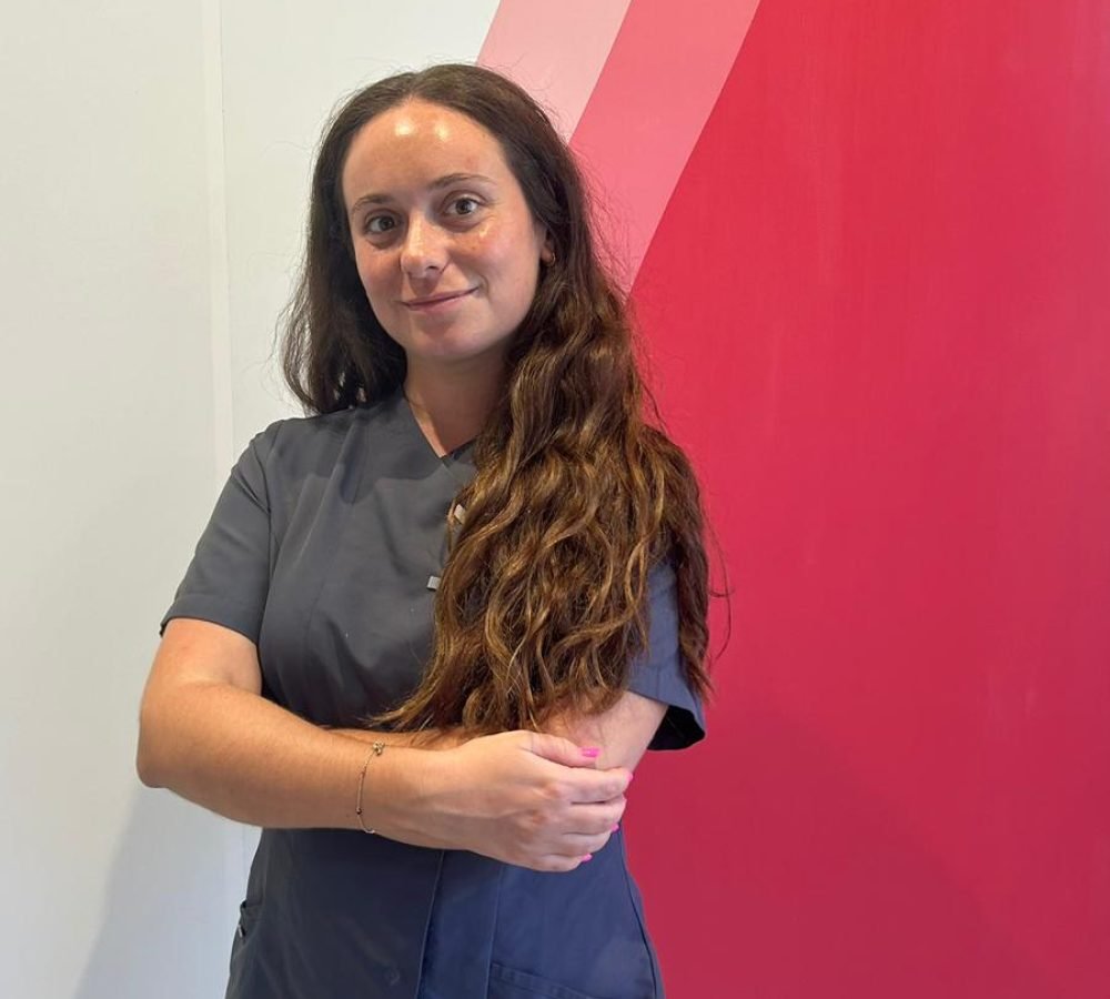 Fotografía de la Dra. Pilar Moreno Palma, odontóloga general, sonriendo con los brazos cruzados. Lleva un uniforme gris y está frente a un fondo blanco con detalles en rojo. El texto indica su nombre y especialidad en cirugía, periodoncia e implantes.