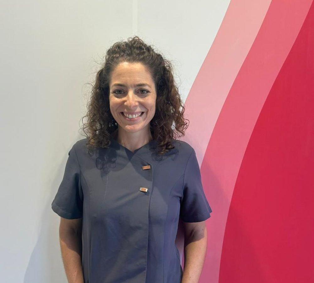 Fotografía de la Dra. Elena García Ocaña, odontóloga colegiada, sonriente y con los brazos cruzados. Lleva un uniforme gris y está de pie frente a un fondo blanco con detalles en rojo, donde se puede leer su nombre y especialidad en estética dental.